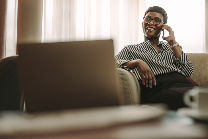 Businesswoman relaxing on hotel room couch while talking on phone and working on laptop during business trip
